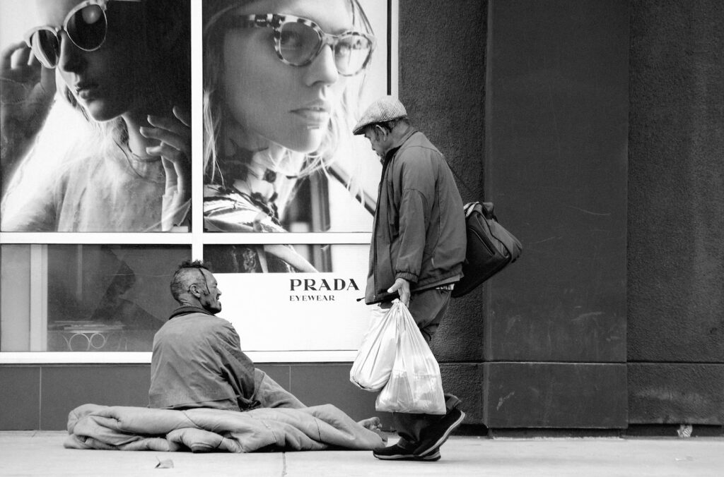 grayscale photo of man with shop bags walking past beggar siting on sidewalk